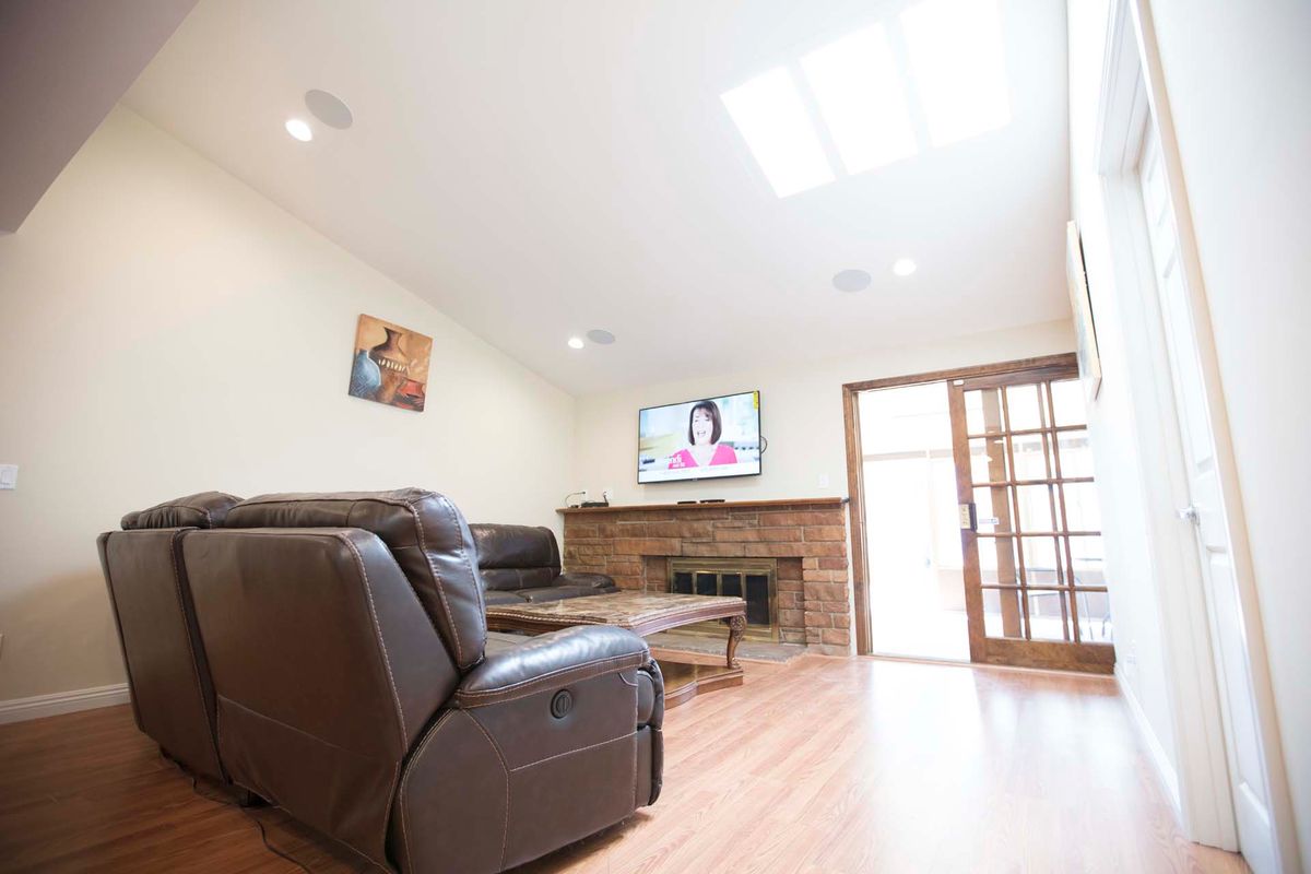 A spacious living room with a leather sofa, fireplace, and a wall-mounted TV. The room is brightened by a skylight and features wooden flooring.