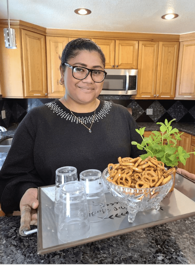 A person holding a tray with glasses, pretzels, and a small plant in a kitchen.