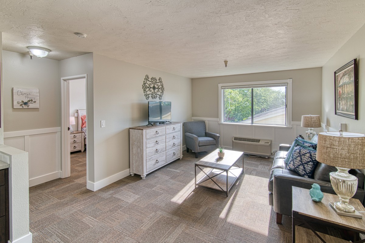 A bright and spacious living room with a sofa, armchair, and a coffee table in front of a TV.