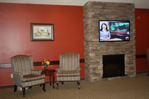 A cozy living room with a stone fireplace, a wall-mounted television, and two striped chairs beside a small table with flowers.