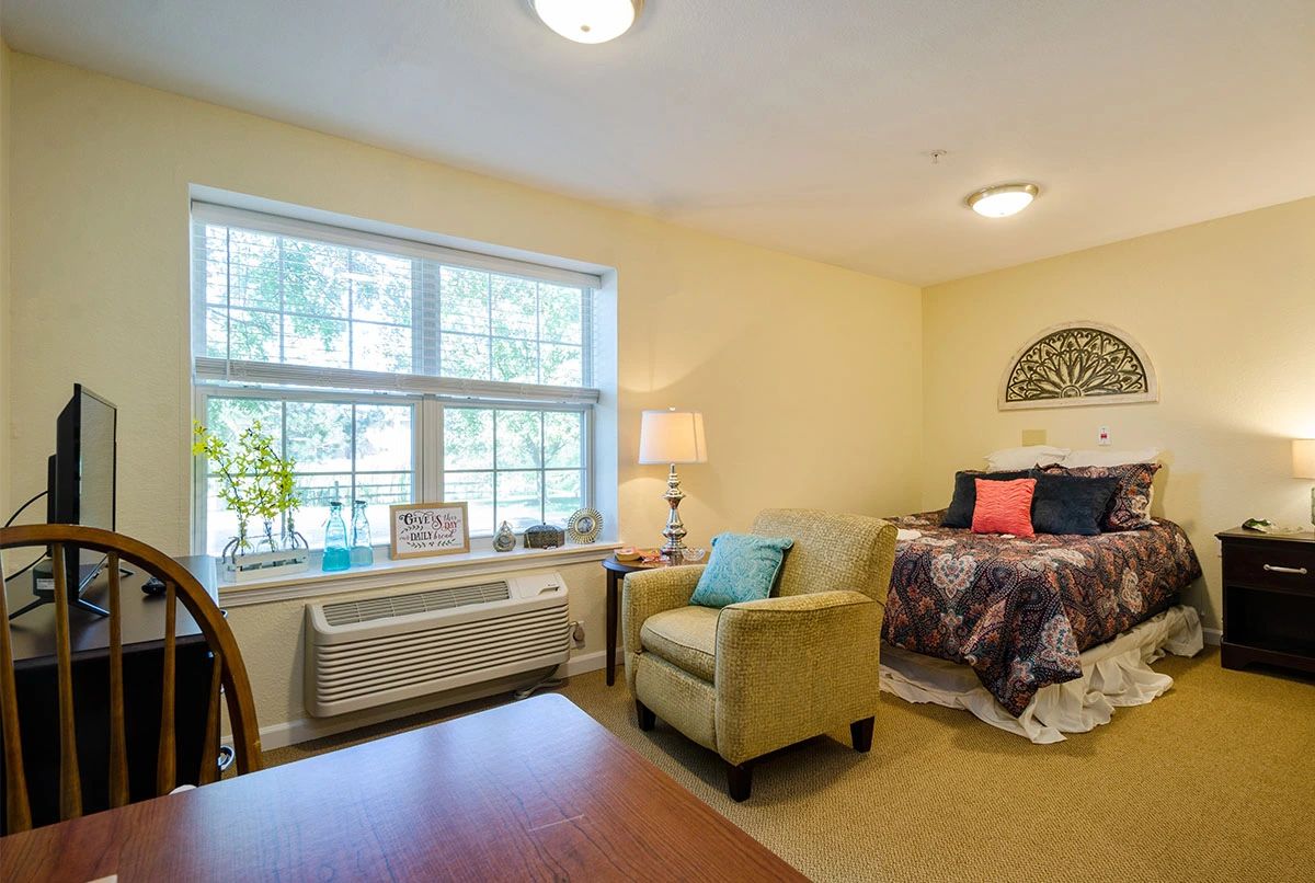 A cozy bedroom with a bed, armchair, and large window letting in natural light.