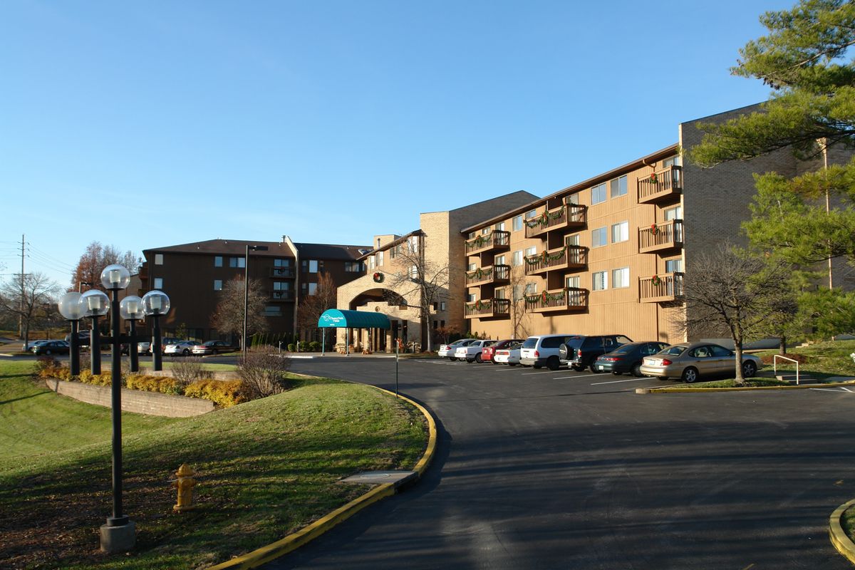 A four-story hotel building with a parking lot in front, under a clear blue sky.