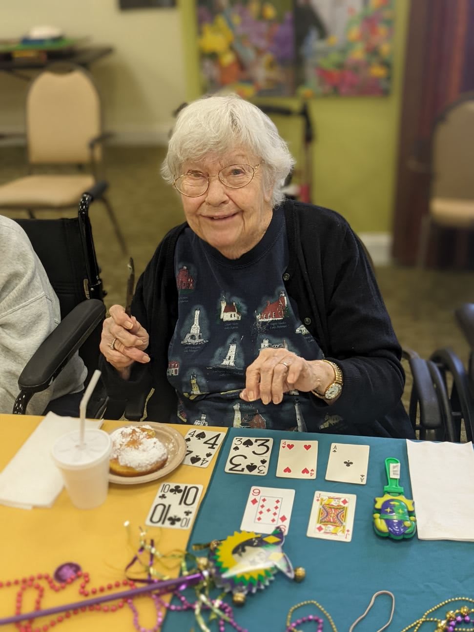 An elderly woman smiling while playing a game of cards at a table indoors.