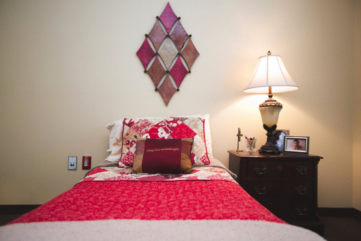 A cozy bedroom with red bedding and a bedside lamp, featuring decorative wall art.
