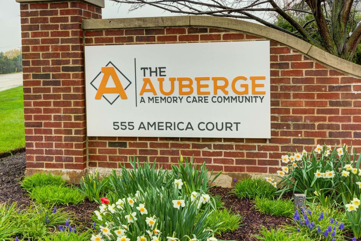 Sign for The Auberge, a memory care community, at 555 America Court surrounded by landscaped flowers.