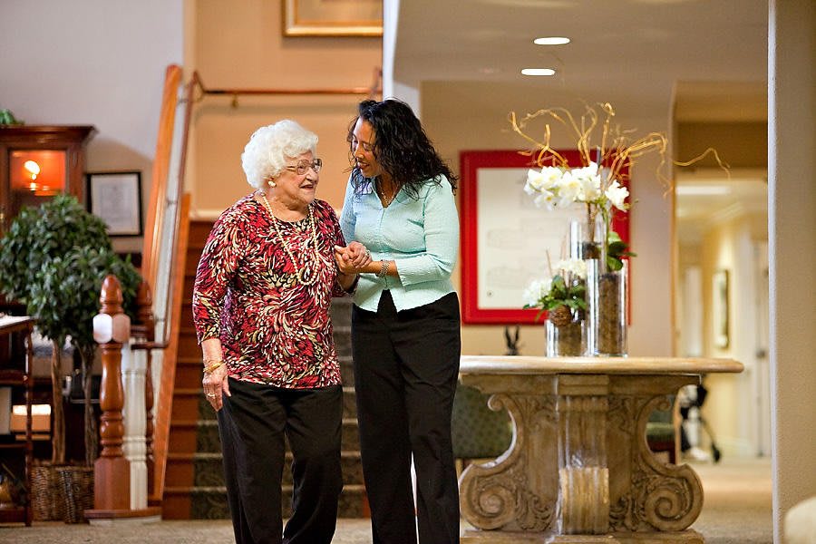 A caregiver assists an elderly woman, both smiling warmly as they walk together indoors.