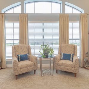 A cozy living room setup featuring two armchairs and a small glass coffee table in front of large windows with curtains.