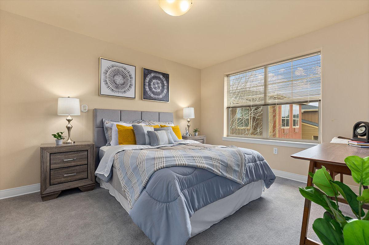 A cozy bedroom featuring a neatly made bed with blue and yellow accents, flanked by two bedside tables and lamps, with decorative artwork on the walls and a large window providing natural light.