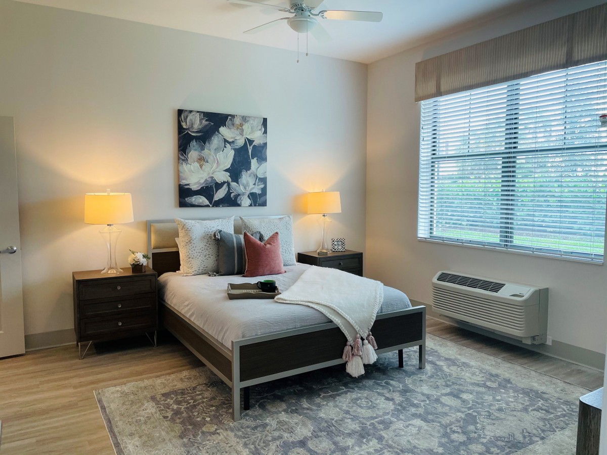 A cozy bedroom with a neatly made bed, bedside lamps, a large window, and decorative painting on the wall.