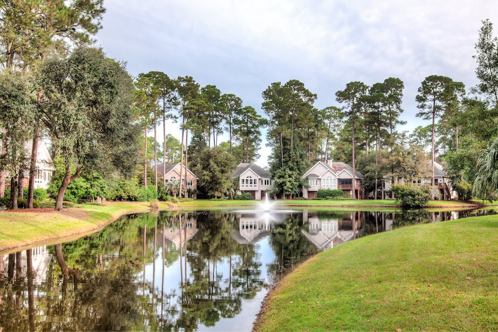 A scenic view of houses by a tranquil lake with a fountain and surrounding trees.