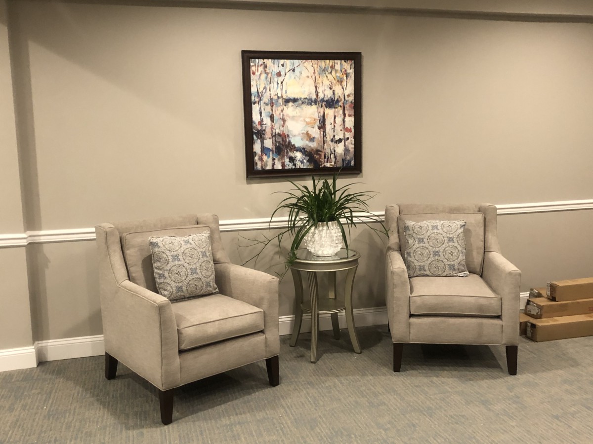 Two beige armchairs with cushions, a small side table with a plant, and framed artwork on the wall create a cozy living room setting.