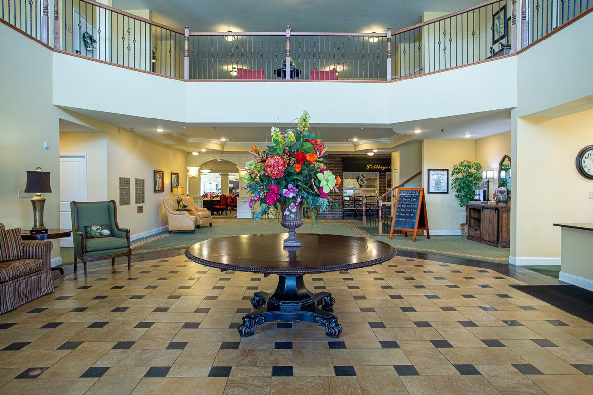 A spacious lobby featuring a central table with a floral arrangement, surrounded by elegant decor and an upper balcony.