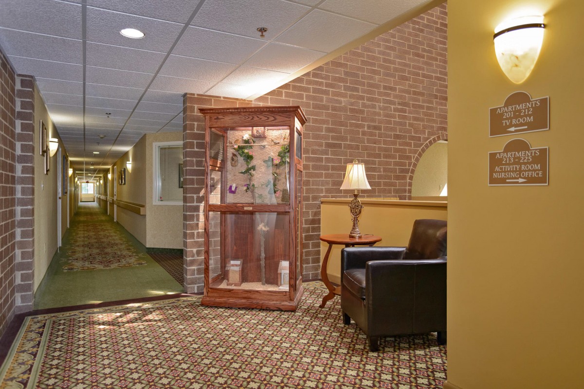 A cozy hallway with a brick wall, featuring a display case, a leather chair, and a lamp on a side table.