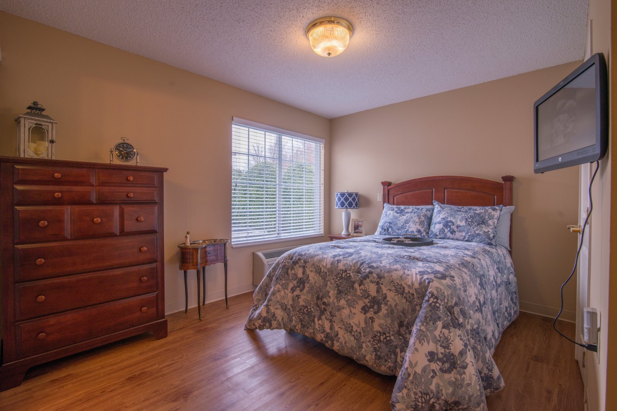 A cozy bedroom with a wooden floor, featuring a double bed, dresser, and a window allowing natural light.