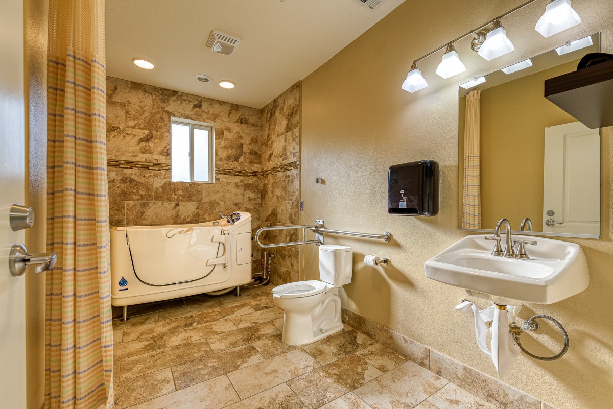 An accessible bathroom featuring a walk-in tub, toilet, sink, and grab bars with tile flooring.