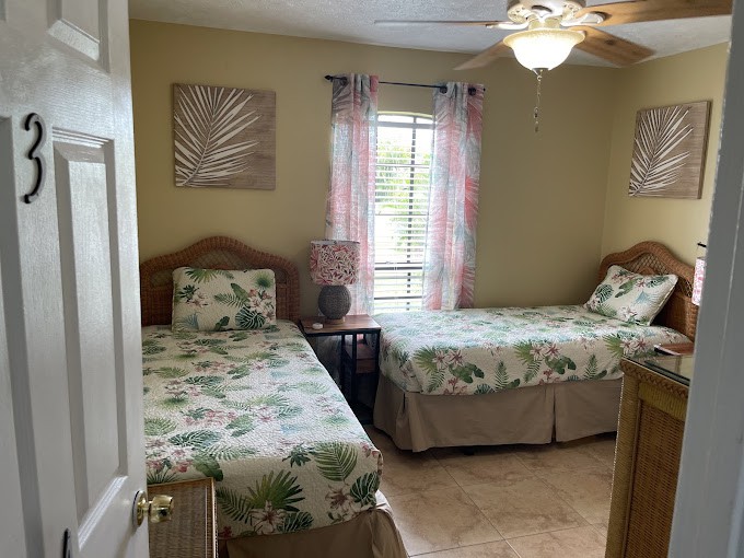 A cozy bedroom with two twin beds featuring floral bedding, complemented by a ceiling fan and soft natural light from the window.