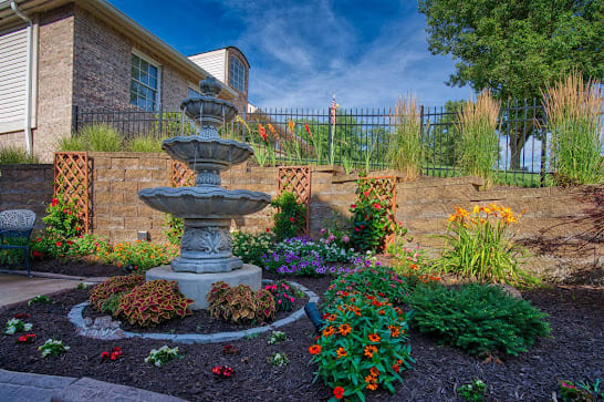 A beautifully landscaped garden featuring a multi-tiered fountain surrounded by vibrant flowers and a brick retaining wall.