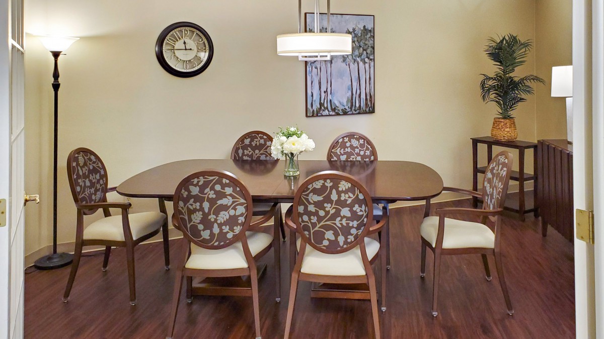 A cozy dining room with a wooden table and floral pattern chairs, illuminated by a pendant light, featuring wall art and a clock.