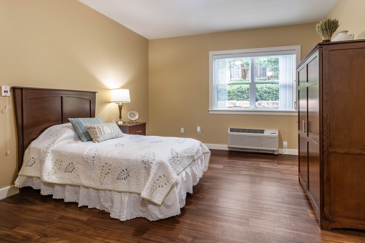 A cozy bedroom with a bed, nightstand, lamp, and a large window letting in natural light.