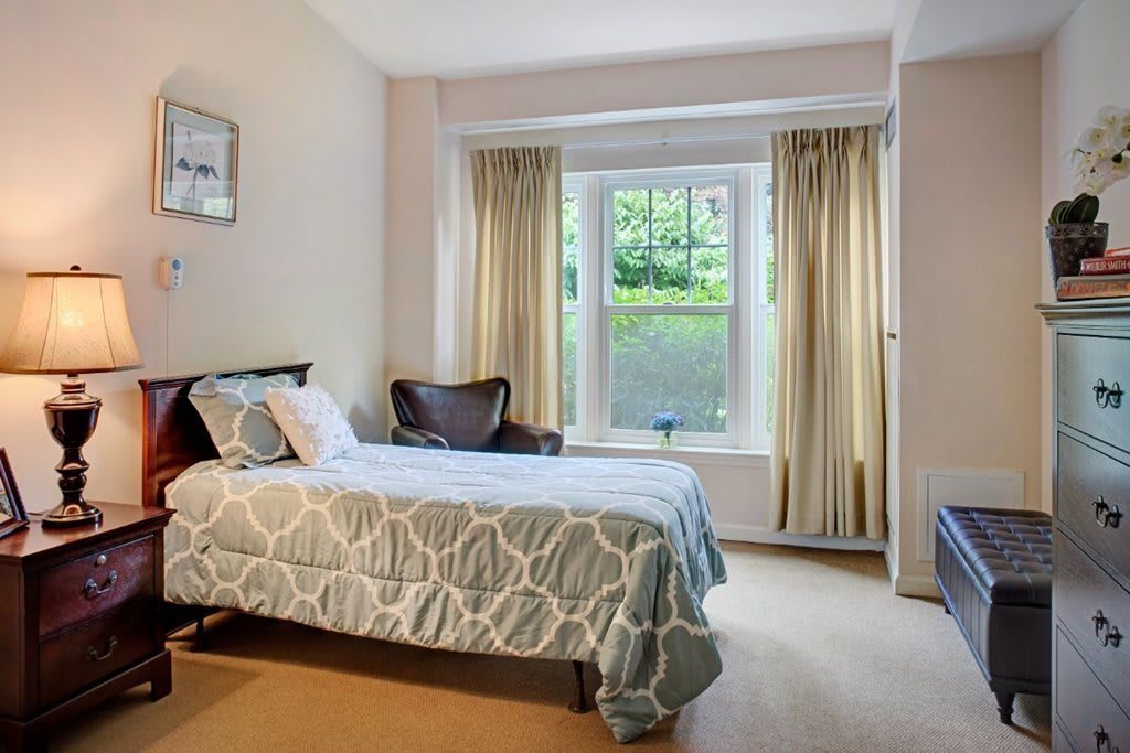 A cozy bedroom featuring a single bed with patterned bedding, a large window with curtains, and natural daylight.