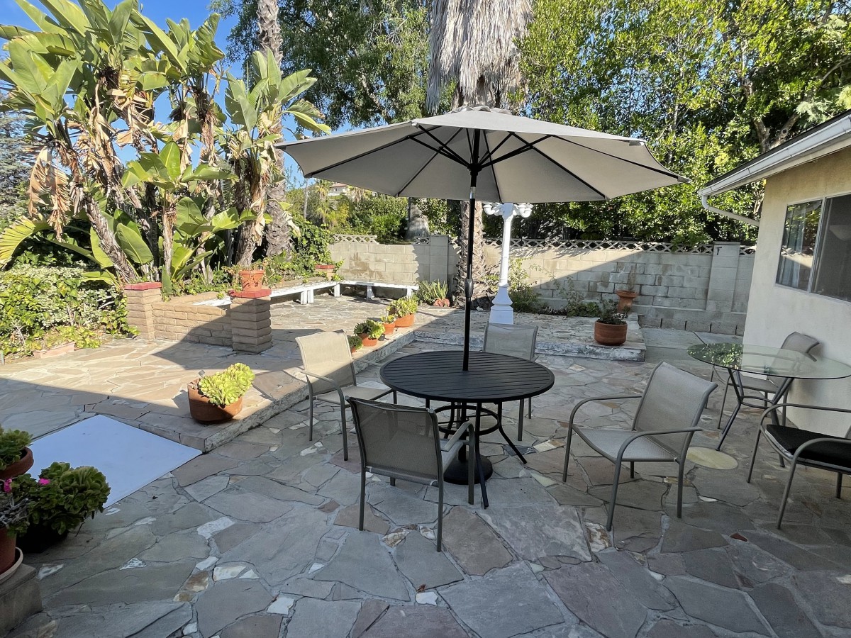 A sunny patio with outdoor seating and a large umbrella, surrounded by lush greenery.