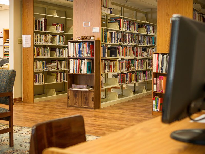 A quiet library setting with bookshelves filled with various books and a computer workstation in the foreground.