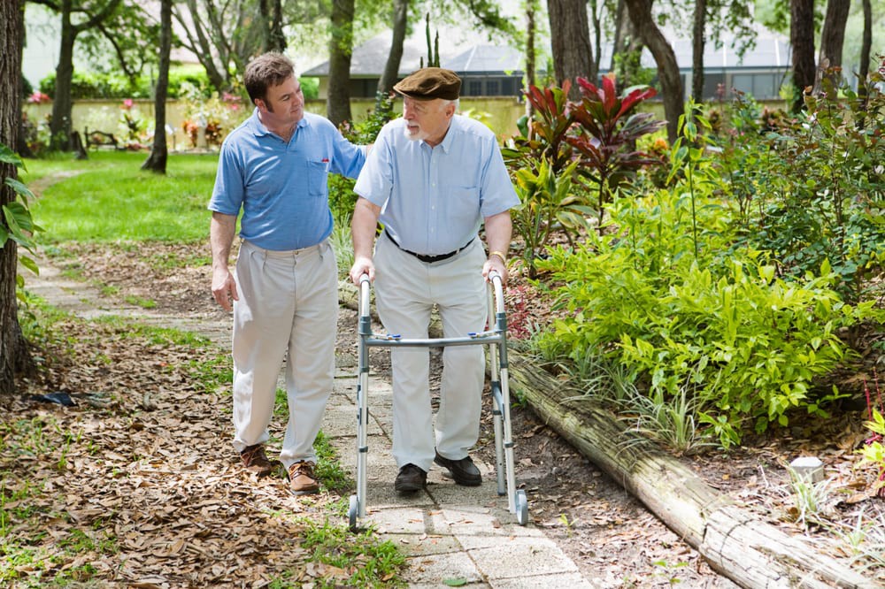 An elderly man using a walker is assisted by a caregiver as they walk together in a park.
