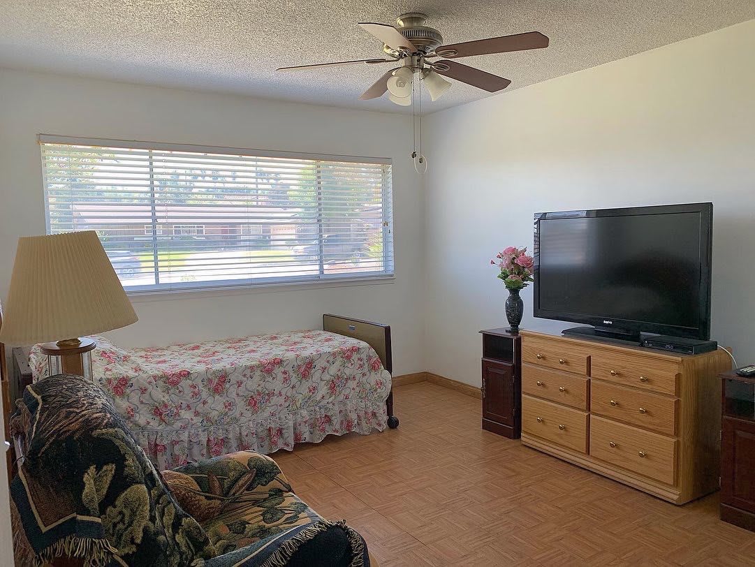 A cozy bedroom with a single bed, dresser, and television, featuring a large window and ceiling fan.