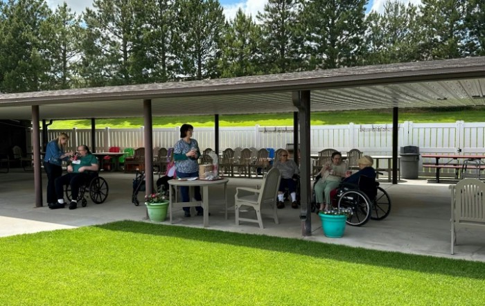 A group of people gathered under a covered patio with tables and chairs, surrounded by greenery.