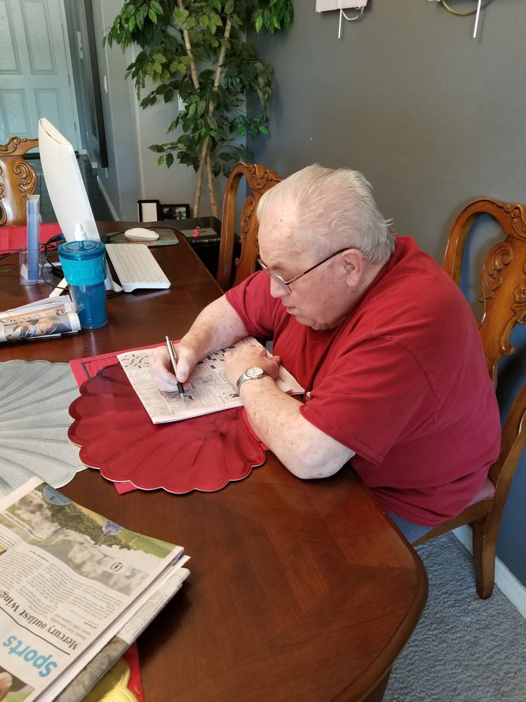 An elderly man in a red shirt is writing at a table indoors.