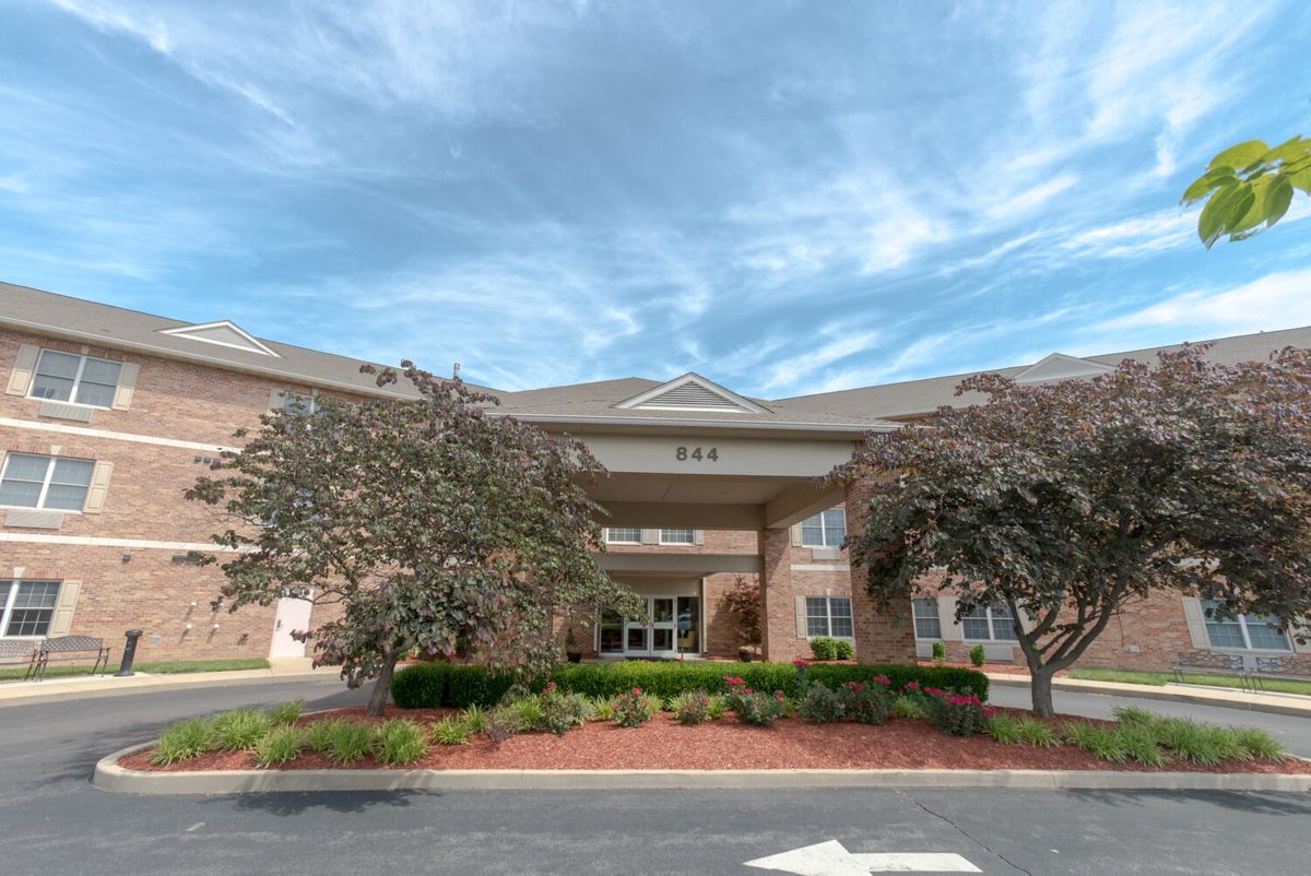 The front entrance of a brick building surrounded by trees and landscaping under a cloudy sky.