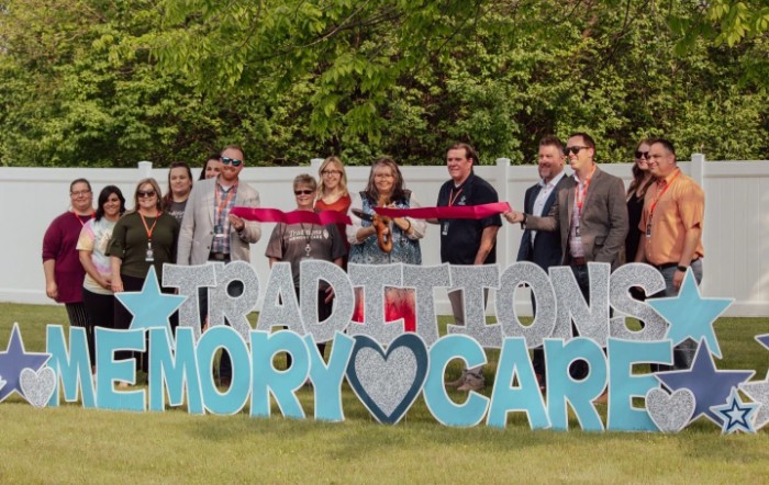 A group of people participating in a ribbon-cutting ceremony for a memory care facility outdoors.