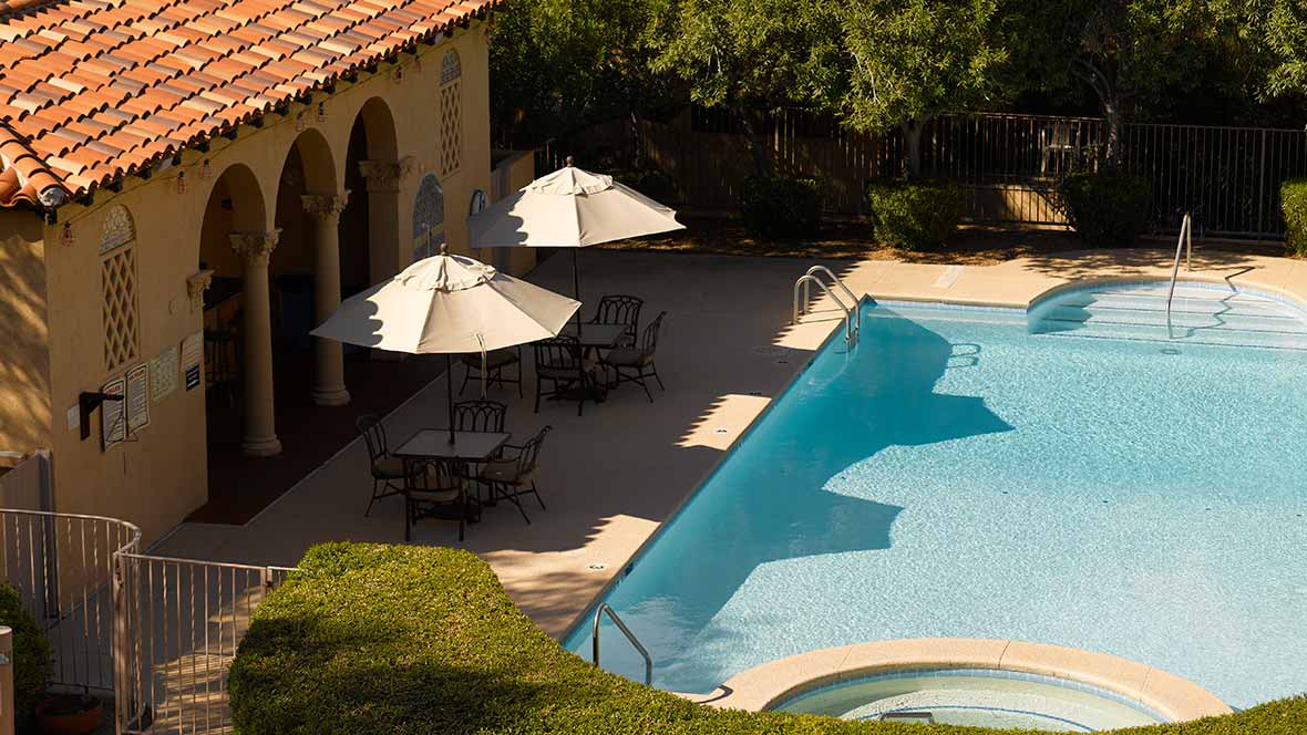 A serene outdoor setting featuring a swimming pool alongside shaded tables and chairs, with trees in the background.