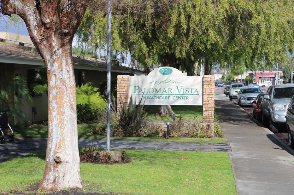 Entrance sign for an apartment complex surrounded by trees with a nearby sidewalk and parked cars.