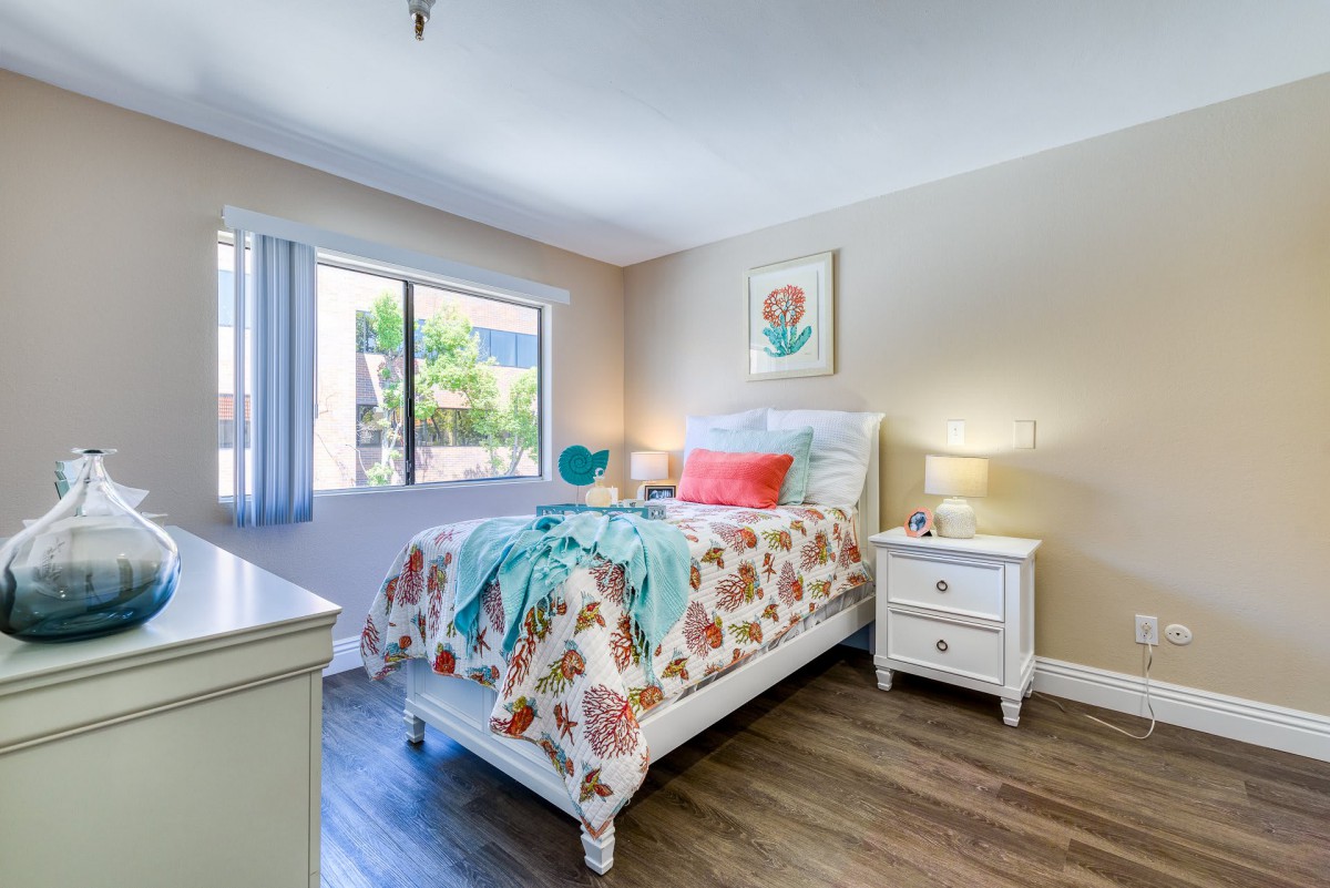 A bright and cozy bedroom with floral bedding, a window allowing natural light, and a bedside table adorned with a lamp.
