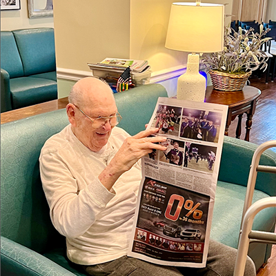 An elderly man sits comfortably in a chair, reading a newspaper under the warm glow of a lamp.