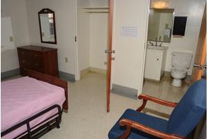 A simple room with a bed, chair, mirror, open closet, and a sink area.