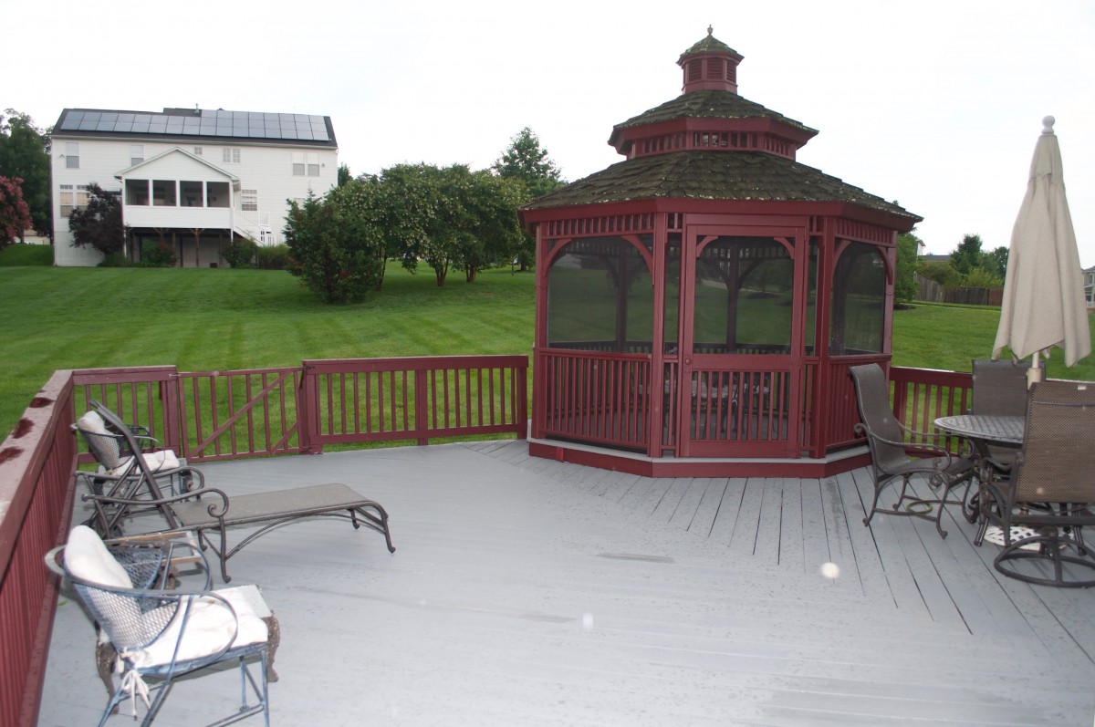 A wooden gazebo on a spacious deck with outdoor furniture, overlooking a green lawn.