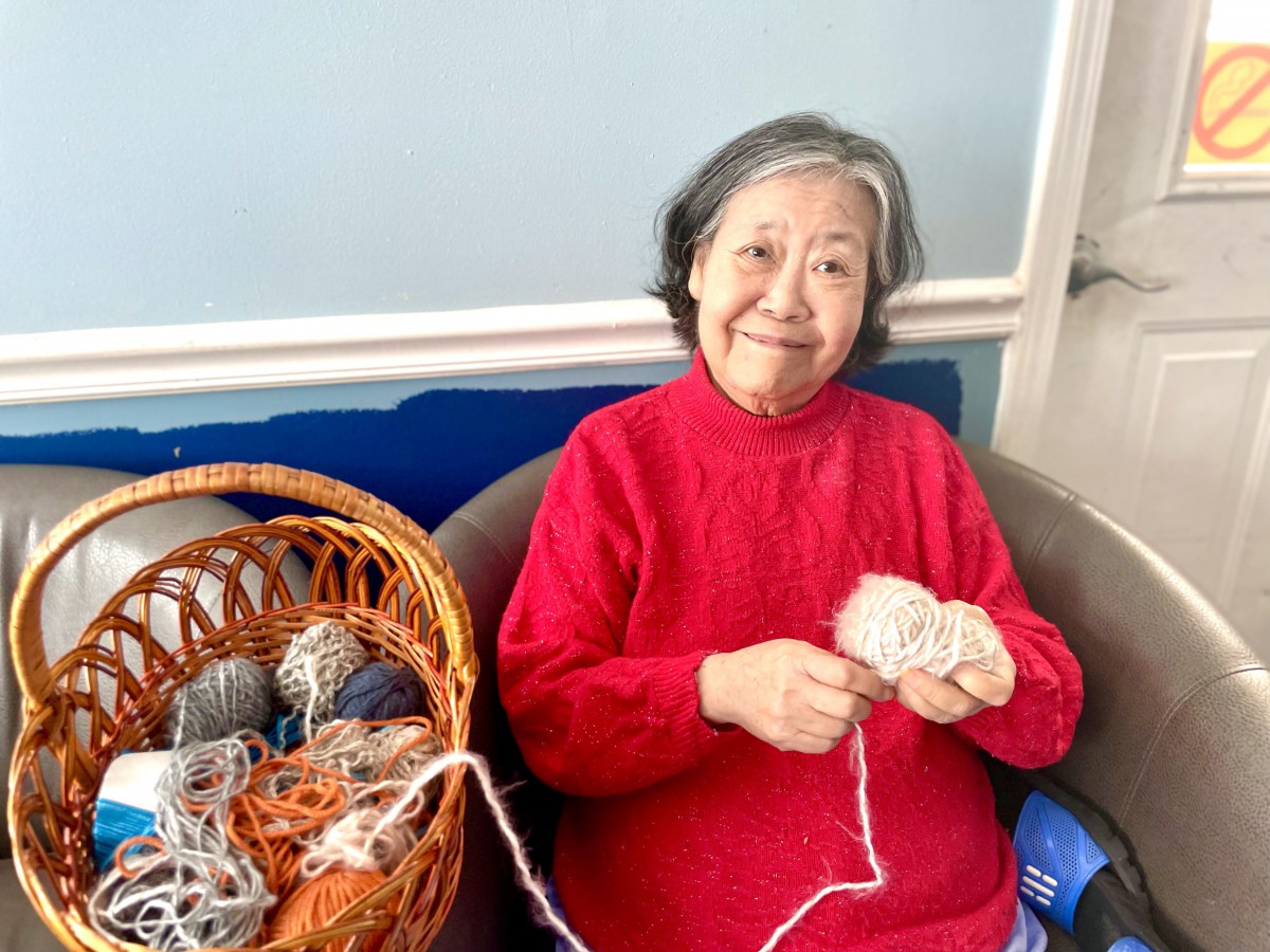An elderly woman knitting with yarn next to a basket filled with colorful yarn balls.