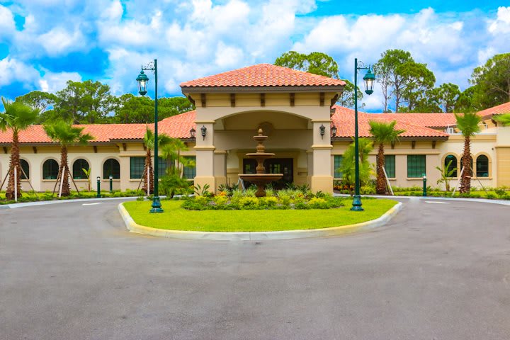 A scenic view of a Mediterranean-style building with a central fountain and surrounding greenery under a partly cloudy sky.