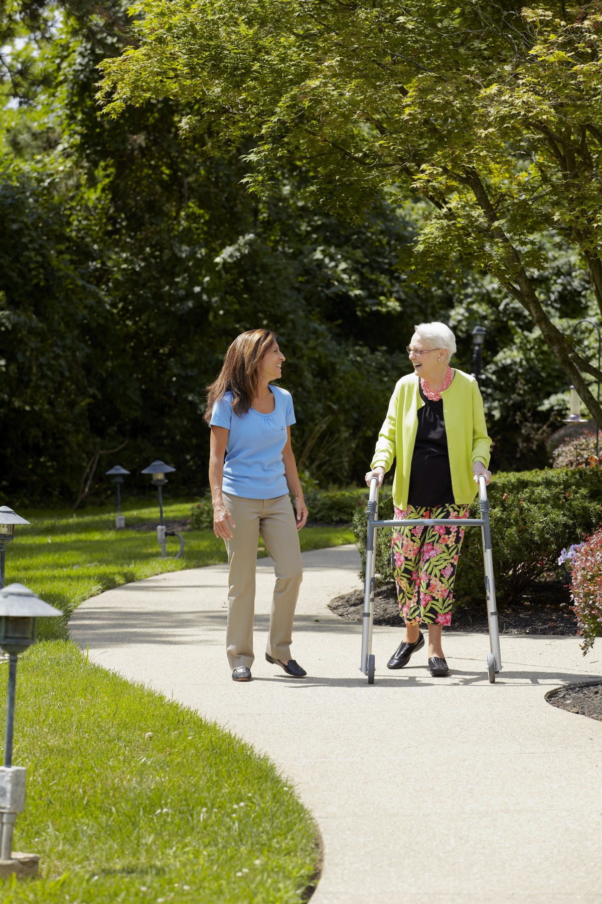 An elderly woman with a walker is accompanied by a caregiver as they walk along a garden path.