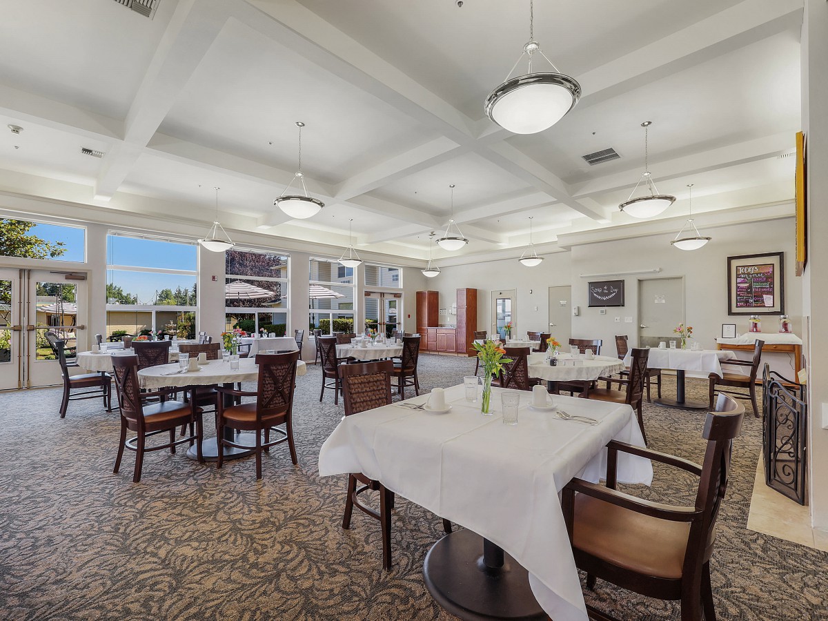 A spacious dining room with elegantly set tables, white tablecloths, and large windows allowing natural light.