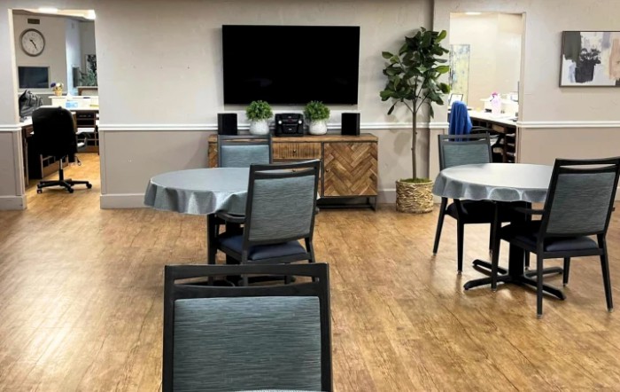 A cozy room with tables and chairs, featuring a television on the wall and decorative plants.