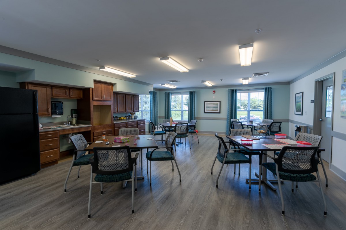 A community dining room with multiple tables and chairs, featuring a kitchen area and large windows.