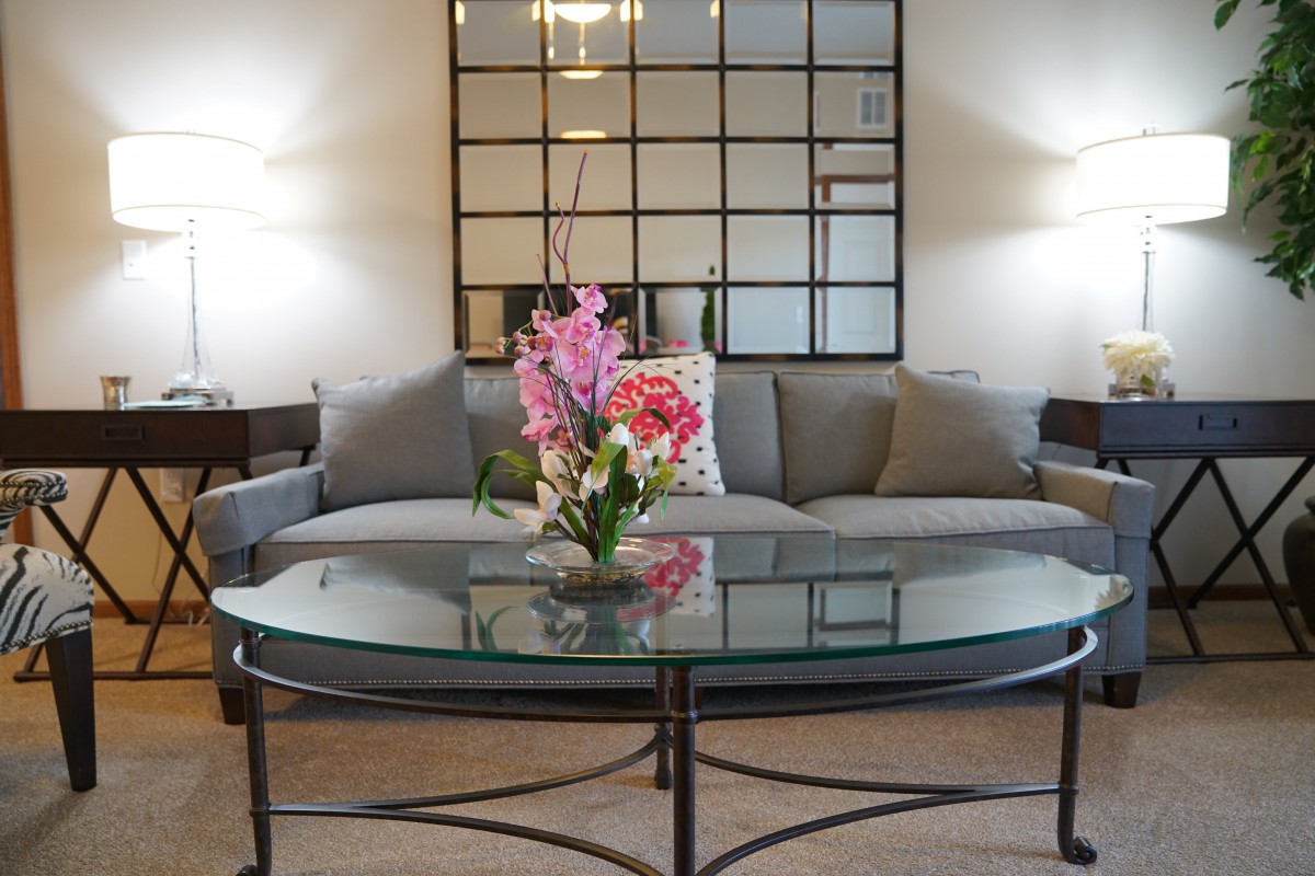 A modern living room featuring a gray sofa, glass coffee table with flowers, and a mirrored wall.