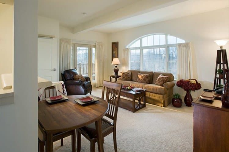 A cozy living room with a sofa, armchair, dining table, and large windows.