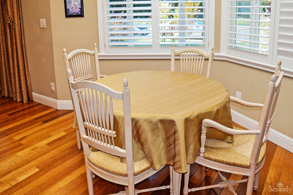 A cozy dining area with a round table covered by a beige tablecloth, surrounded by four chairs, and natural light streaming through the window blinds.