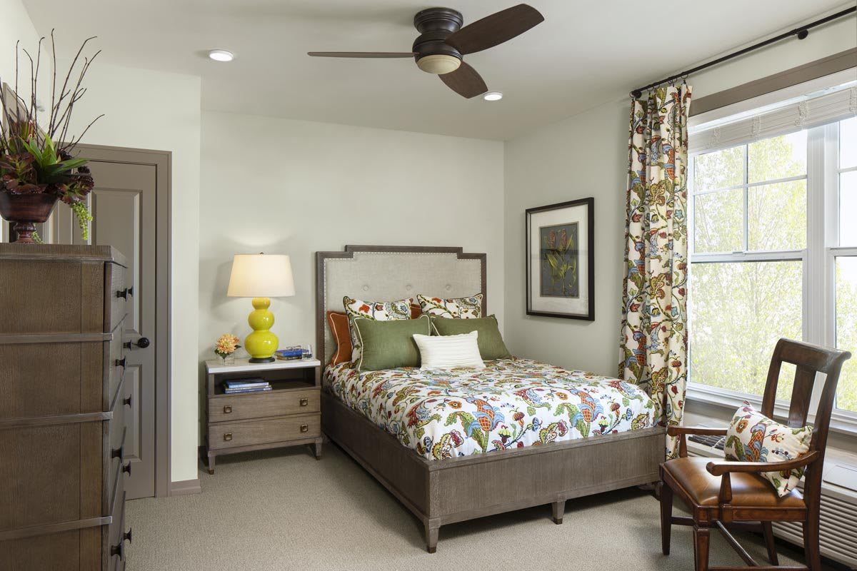 A cozy bedroom featuring a comfortable bed with floral bedding, a nightstand with a lamp, and a ceiling fan.