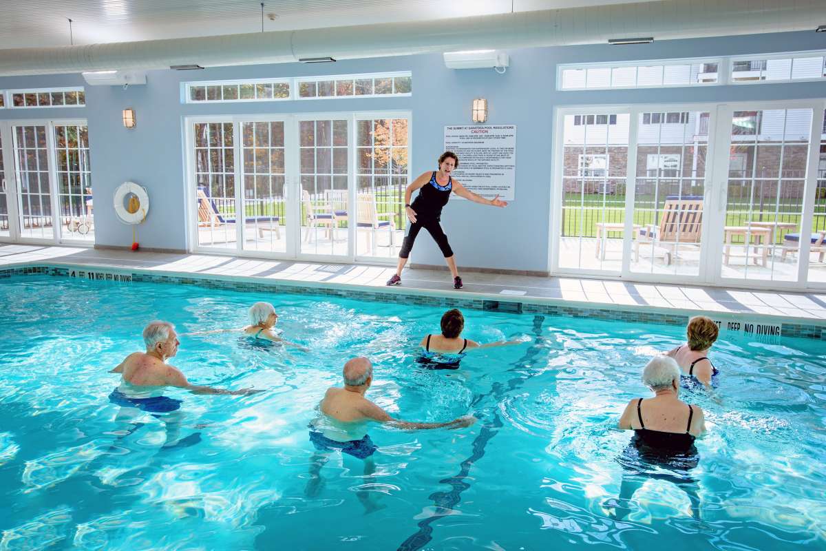 A group of seniors participating in a water aerobics class led by an instructor at an indoor pool.
