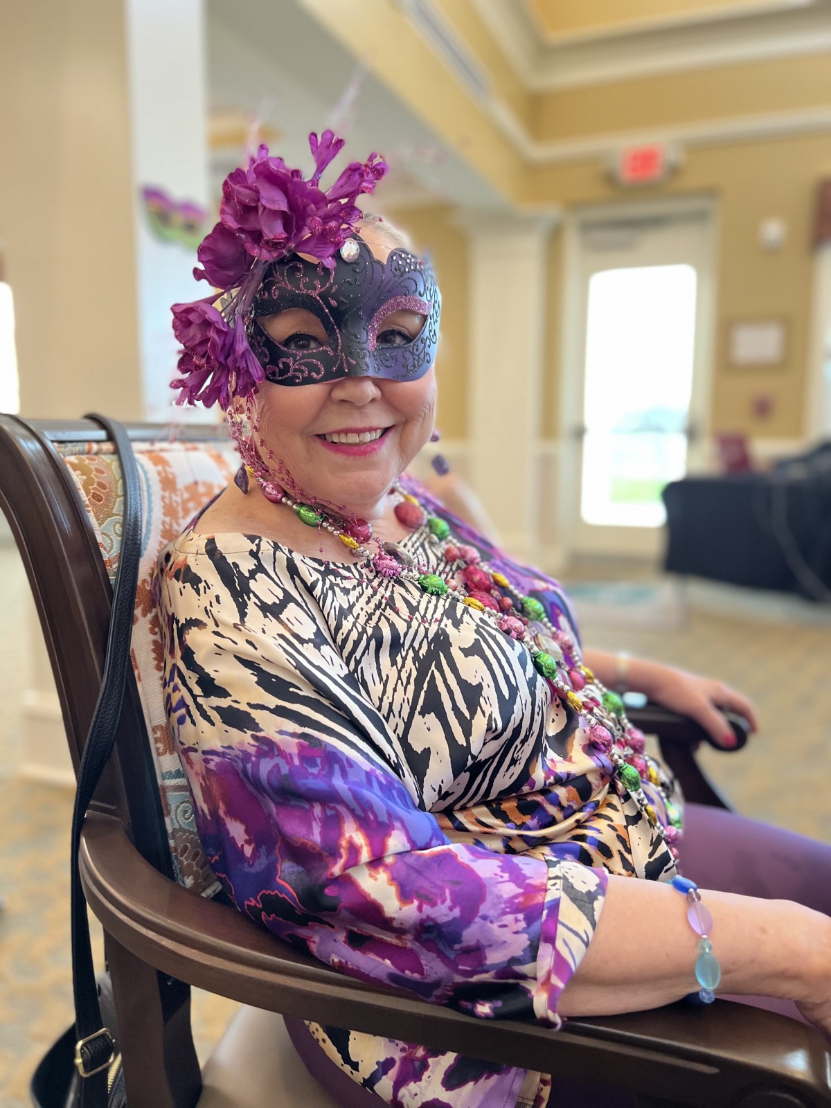 A person wearing a decorative mask and floral accessories, seated indoors.